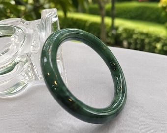 May include: A smooth, circular bangle bracelet crafted from dark green stone with subtle, lighter green marbled veining throughout. The bangle features a polished, glossy finish that reflects light, showcasing a deep, rich color profile and a solid, continuous band structure.