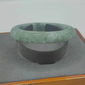Könnte beinhalten: Ein hellgrünes Jade-Armreif-Armband mit geschnitzten Blumenmustern. Das Armband wird auf einem grauen Ständer in einer Holzkiste präsentiert. Der Jade hat eine durchscheinende Qualität, und die Schnitzereien verleihen dem Stück Textur und Detail.