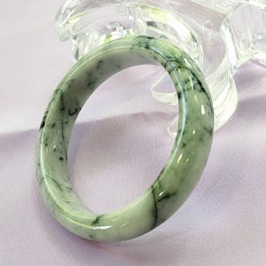 May include: A jade bangle bracelet with a smooth, polished surface. The bracelet is a light green color with dark green veining throughout. The bangle is round and appears to be made of a solid piece of jade.