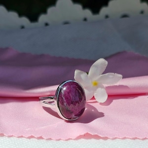 Puede incluir: Un anillo de plata con una gran piedra preciosa ovalada de color púrpura engastada en el centro. El anillo está sobre un fondo de tela rosa con una flor blanca.