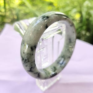 May include: A jade bangle bracelet with a marbled appearance. The bracelet is primarily light green with dark green and black veining throughout. The smooth, polished surface reflects light, highlighting the natural patterns. The bracelet is displayed on a clear stand.