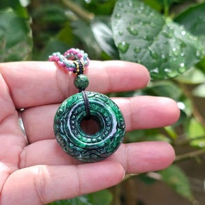 May include: A dark green jade pendant with intricate carved details. The circular pendant is suspended from a black cord and a colorful braided necklace. The jade has a polished finish and a central hole.