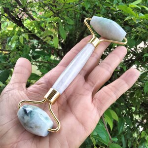 Op de afbeelding: Een jade gezichtsroller met een witte en gouden handgreep. De roller heeft twee jadestenen, één aan elk uiteinde, verbonden door goudkleurig metaal. De jadestenen zijn lichtgroen van kleur met donkerdere groene adering.