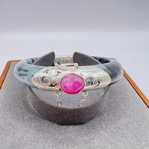 May include: A jade bangle bracelet with a silver clasp and a large, round, pink gemstone. The jade is a mix of blue and green hues. The silver clasp has an ornate design.