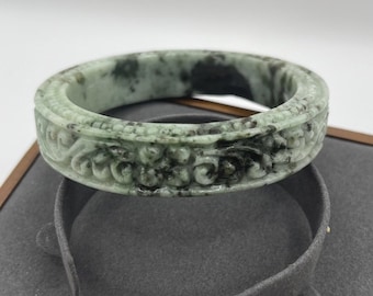 May include: A green jade bangle with intricate carved floral designs. The bangle has a mottled green and black color pattern.