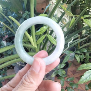 May include: A pale green jade bangle bracelet. The circular bracelet is held in front of a background of green foliage. The jade has a smooth, polished surface and a translucent appearance.