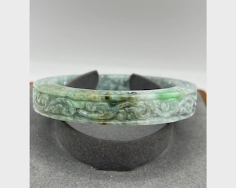May include: A jade bangle bracelet with intricate carved floral patterns. The bracelet is a mix of green hues, with darker green accents. The design is visible on the outer surface of the bracelet. The bracelet is displayed on a black stand.