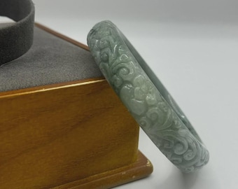 May include: A pale green jade bangle bracelet with intricate floral and scroll carvings. The bracelet is displayed on a wooden box with a gray interior, showcasing the detailed craftsmanship of the jewelry.