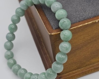 May include: A light green beaded bracelet. The bracelet is made of round, polished beads with a subtle, translucent quality. The beads are strung together, forming a circular shape. The bracelet is resting on a wooden surface.