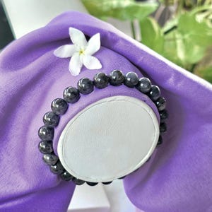 May include: A dark gray beaded bracelet with a polished finish. The bracelet is displayed on a white oval-shaped stand, set against a backdrop of a purple fabric and a white flower. The beads appear to be approximately 0.3 inches in diameter.