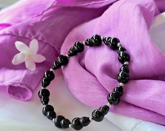 May include: A black beaded bracelet with a unique design, resting on a purple scarf. The bracelet features glossy, rounded beads connected by small silver-colored links. A white flower is placed near the bracelet.