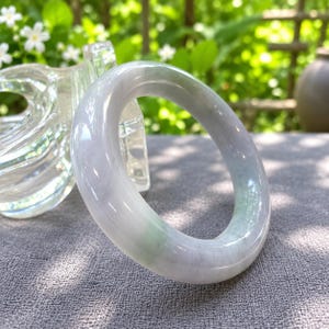 May include: A smooth, polished bangle bracelet crafted from translucent stone, featuring a rounded profile with subtle, blended hues of pale lavender, soft white, and faint green streaks throughout the circular band's polished surface.