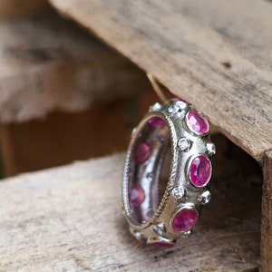 Puede incluir: Un anillo de plata con piedras preciosas de rubí de corte ovalado y pequeños diamantes redondos. El anillo tiene una banda texturizada. Los rubíes son de color rojo vibrante. El fondo es de madera.