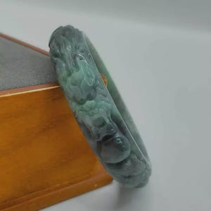 Puede incluir: Un brazalete de jade con un diseño de dragón tallado. El brazalete es una mezcla de tonos verdes y grises, con los detalles del dragón intrincadamente detallados. El brazalete se exhibe sobre una superficie de madera.