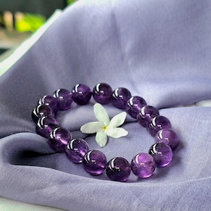 Può includere: Un braccialetto con perline rotonde di ametista lucida, di colore viola intenso. Le perline sono disposte a cerchio, con un fiore bianco al centro. Il braccialetto è appoggiato su un tessuto lavanda.