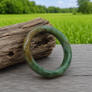 May include: A jade bangle bracelet with a smooth, polished surface. The bracelet features a blend of green and brown hues, with a translucent quality. The bangle is displayed on a piece of weathered wood against a backdrop of green foliage.
