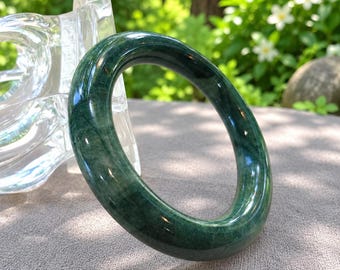May include: A smooth, polished bangle bracelet crafted from dark green stone with visible natural veining and mottled patterns throughout the circular band. The surface of the bracelet has a high-gloss finish that reflects light, showcasing the deep forest green color and the intricate, organic mineral inclusions within the solid stone structure.
