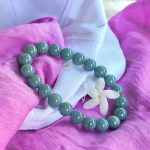 May include: A jade bead bracelet with a soft, seafoam green hue. The bracelet is composed of round, polished beads. A white flower and pink fabric provide a contrasting backdrop.