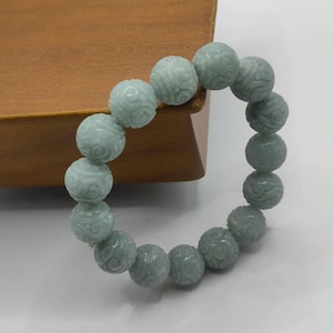 Op de afbeelding: Een lichtgroene kralenarmband. De armband heeft ronde kralen met gesneden bloempatronen. De kralen zijn aan elkaar geregen om een cirkelvorm te vormen. De armband wordt getoond op een wit oppervlak met een houten achtergrond.