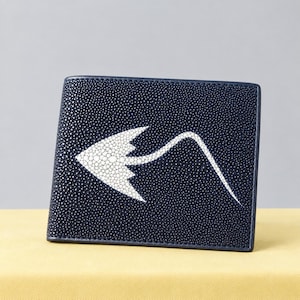 Könnte beinhalten: Ein dunkelblaues Bifold-Portemonnaie mit einer texturierten, genarbten Oberfläche. Auf der Vorderseite befindet sich ein markantes, eingelegtes weißes Design in Form eines Stachelrochens mit langem, geschwungenem Schwanz aus Kontrastmaterial.