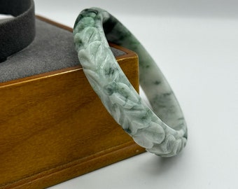May include: A carved jade bangle with a floral design. The bangle is a light green color with white marbling.
