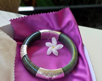 May include: A green jade bangle bracelet with silver accents and a floral design. The bracelet is on a purple satin cloth with a white flower.