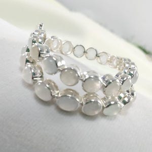 Op de afbeelding: Twee zilveren ringen met ovale, melkachtig witte edelstenen. De ringen zijn op een witte ondergrond gestapeld. De edelstenen zijn in een doorlopend patroon rond de ringen geplaatst. Waarschijnlijk van sterling zilver.