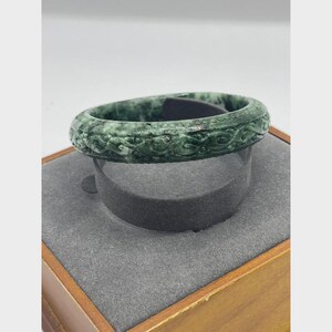 May include: A green jade bangle bracelet with intricate carved details. The bracelet is displayed on a gray stand, showcasing its polished surface and detailed design. The jade's color varies from light to dark green.