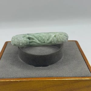 Könnte beinhalten: Ein hellgrünes Jade-Armreif-Armband mit geschnitzten Blumen- und Blattmustern. Das Armband wird auf einem grauen Samtringhalter in einer Holzkiste präsentiert. Der Jade hat eine durchscheinende Qualität.