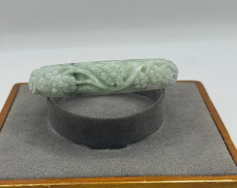 May include: A light green jade bangle bracelet with carved floral and leaf designs. The bracelet is displayed on a gray velvet ring holder inside a wooden box. The jade has a translucent quality.