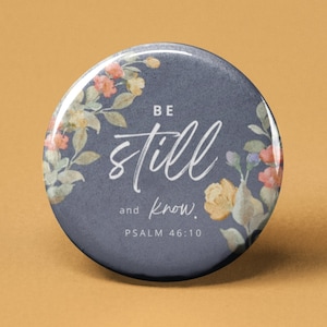 May include: A round button with a blue background and a floral design. The text "BE still and know. PSALM 46:10" is written in white.
