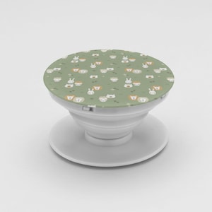 May include: A white phone grip with a round, green top. The top features a repeating pattern of cartoon animal faces, including rabbits and lions. The grip is designed to attach to the back of a phone for easier handling.