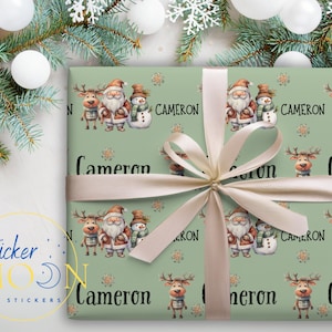 May include: Green wrapping paper with a repeating pattern of reindeer, Santa Claus, and snowmen. The paper is tied with a cream-colored ribbon and the name "Cameron" is printed on the paper.