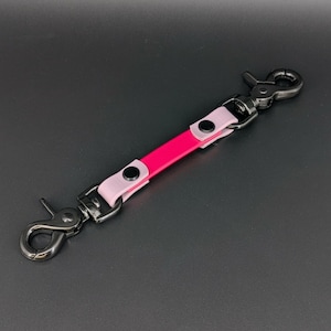 May include: A pink and light pink leather keychain strap with black metal hardware. The strap has two snap hooks and a central pink section. The light pink sections are on either side of the pink section.