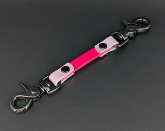 May include: A pink and light pink leather keychain strap with black metal hardware. The strap has two snap hooks and a central pink section. The light pink sections are on either side of the pink section.