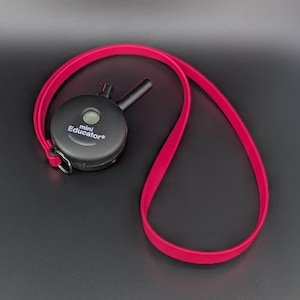 May include: A black Mini Educator device with a black antenna and a bright pink strap. The device has the words "Mini Educator" printed on it. The strap is looped and ready to be worn. The device is designed for dog training.