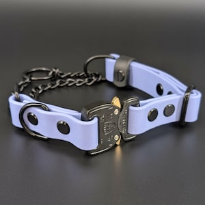 May include: A light purple collar featuring a black metal cobra-style buckle with gold-toned release tabs. The collar includes black metal hardware, including a chain section, D-rings, and rivets securing the strap. The buckle is engraved with text.