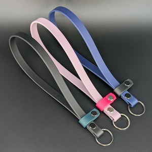 May include: Four leather wrist straps in black, gray, pink, and blue. Each strap has a metal ring and a snap closure. The straps are arranged diagonally against a dark background.