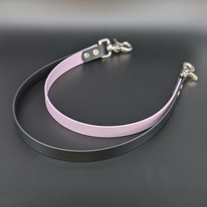 May include: Two leather straps with metal clasps. One strap is black, and the other is light purple. The straps are curved and lie on a dark surface. The clasps are silver-colored.