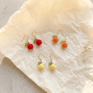 May include: Crochet earrings featuring red, orange, and yellow fruit designs. Each earring has a small crocheted fruit with white flower and green leaf details, hanging from gold-coloured ear wires. The earrings are presented on a cream-coloured paper surface.