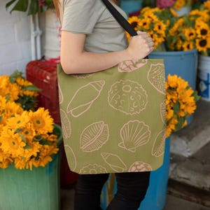 May include: Olive green tote bag with a pink seashell design. The bag has a black strap and is being held in front of a flower shop. The background includes sunflowers and other colourful flowers.