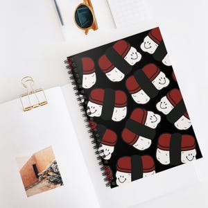 Könnte beinhalten: Ein spiralgebundenes Notizbuch mit schwarzem Einband, der mit einem sich wiederholenden Muster aus lächelnden Sushi-Stücken versehen ist. Das Sushi hat rote, schwarze und weiße Elemente. Das Notizbuch liegt offen auf einer weißen Oberfläche.