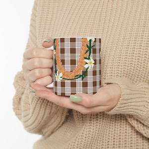 May include: White ceramic mug featuring a brown and white plaid pattern. The mug is decorated with a floral design in shades of orange, white, and green. The mug is held by a person wearing a beige sweater.