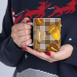 May include: White ceramic mug featuring a repeating pattern of brown and yellow anthurium flowers on a brown plaid background. The mug is held by a person wearing a navy blue sweater with a red reindeer design.