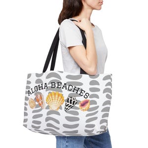 May include: A white tote bag featuring a repeating pattern of grey semi-circular shapes. The bag displays the text ALOHA BEACHES in black letters above a row of five colorful seashells in shades of orange, yellow, white, black, and purple.