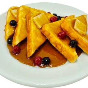 Op de afbeelding: Een bord met goudbruine wentelteefjesdriehoeken, overgoten met siroop, gegarneerd met boter en gegarneerd met verse bosbessen en frambozen. De toast ligt op een wit bord.