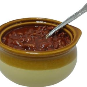 Op de afbeelding: Een keramische kom gevuld met bruine bonen in een donkere, glanzende saus. De kom heeft een lichtbeige onderkant en een bruine bovenkant. Een zilveren lepel ligt in de bonen.