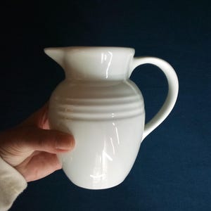 May include: A white ceramic pitcher with a curved handle and a spout. The pitcher has horizontal ridged detailing around the body. The background is a solid dark blue.