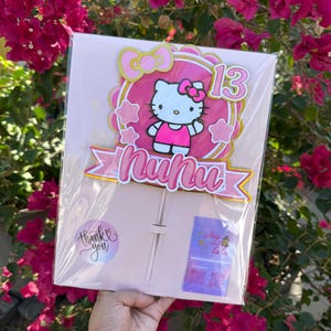 May include: A pink and gold Hello Kitty cake topper with the number 13. The topper features a pink bow, a pink banner with the name "Nunu", and a pink and gold scalloped border. The topper is packaged in a clear plastic bag.
