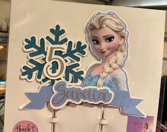 Puede incluir: Adorno para tarta con una imagen de dibujos animados de Elsa de Frozen, un copo de nieve con purpurina verde azulado, el número 5 y el nombre "Saraia" en escritura. El adorno tiene una pancarta azul claro y una pegatina rosa de "gracias".